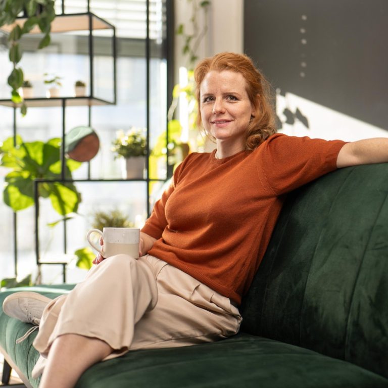 Frau mit rotem Pullover sitzt entspannt auf einer Couch in einem hellen Raum.