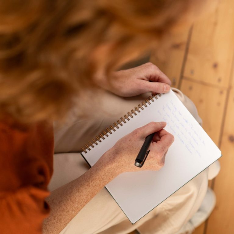 Eine Person in einem roten Pulli schreibt mit einem Stift in ein leeres Notizbuch.