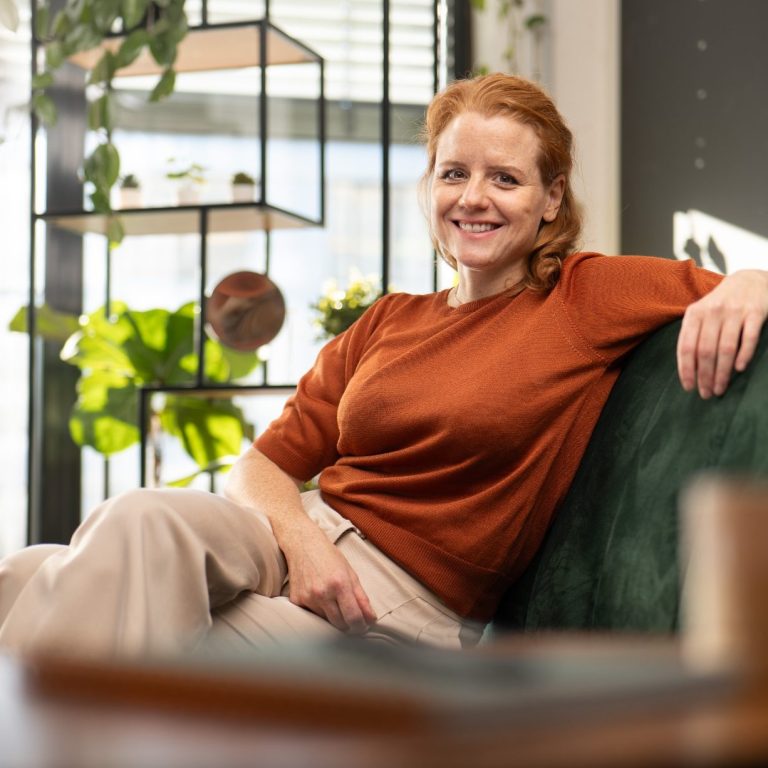 Frau mit rotem Oberteil sitzt lächelnd auf einer Couch in einem hellen Raum.